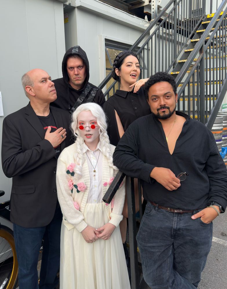 A photo of Jason Carl, Mike Channell, Jane Douglas, Josephine McAdam and Jasper Cartwright posing together before their Vampire the Masquerade Live show in London for World’s Lair. Everyone except for Josephine are dressed in black, while Josephine wears all white with red sunglasses.