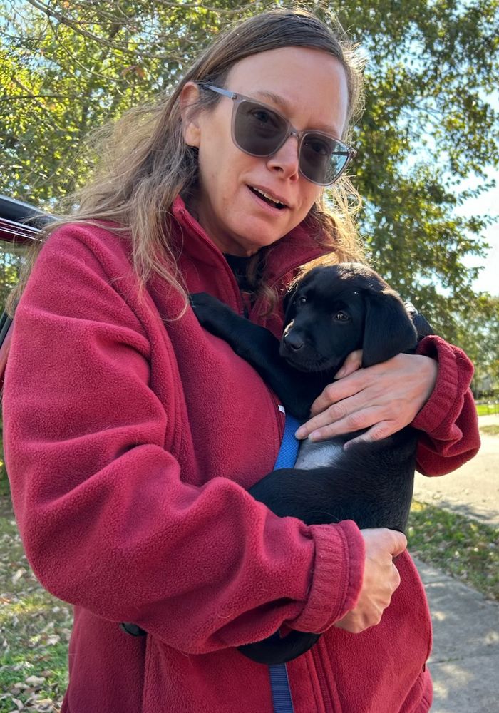 Woman in red jacket holding 6 week old black lab puppy with a blue leash
