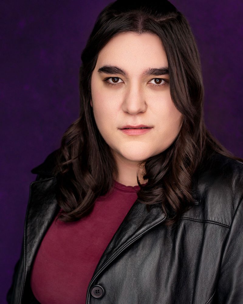 Maren looks deadpan at the camera from a three-quarter view. She wears a maroon shirt and a black leather jacket, with a purple background behind her.