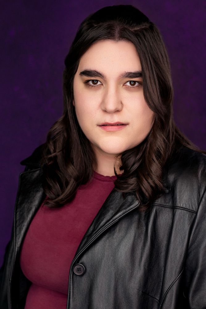 Headshot of Maren looking directly at the camera. She's wearing a black leather jacket, maroon shirt, and stark expression. A dark purple background is behind her.