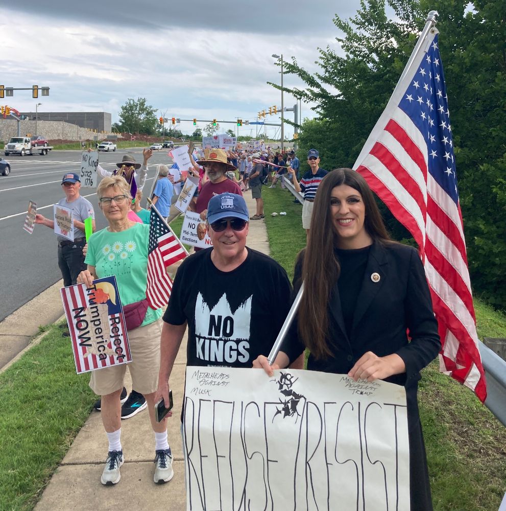 Danica stands on a sidewalk in Haymarket, Virginia with an American flag, a “Refuse/Resist” sign (Sepultura protest song) and a long line of people with signs at the intersection of U.S. 15 and Heathcote Boulevard.