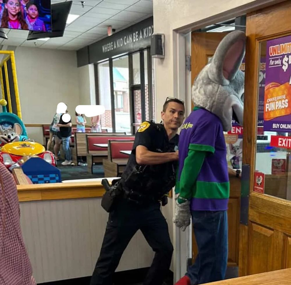 A cop pushing the Chuck E Cheese mascot out the door in front of children because that’s America right now (and Florida daily).
