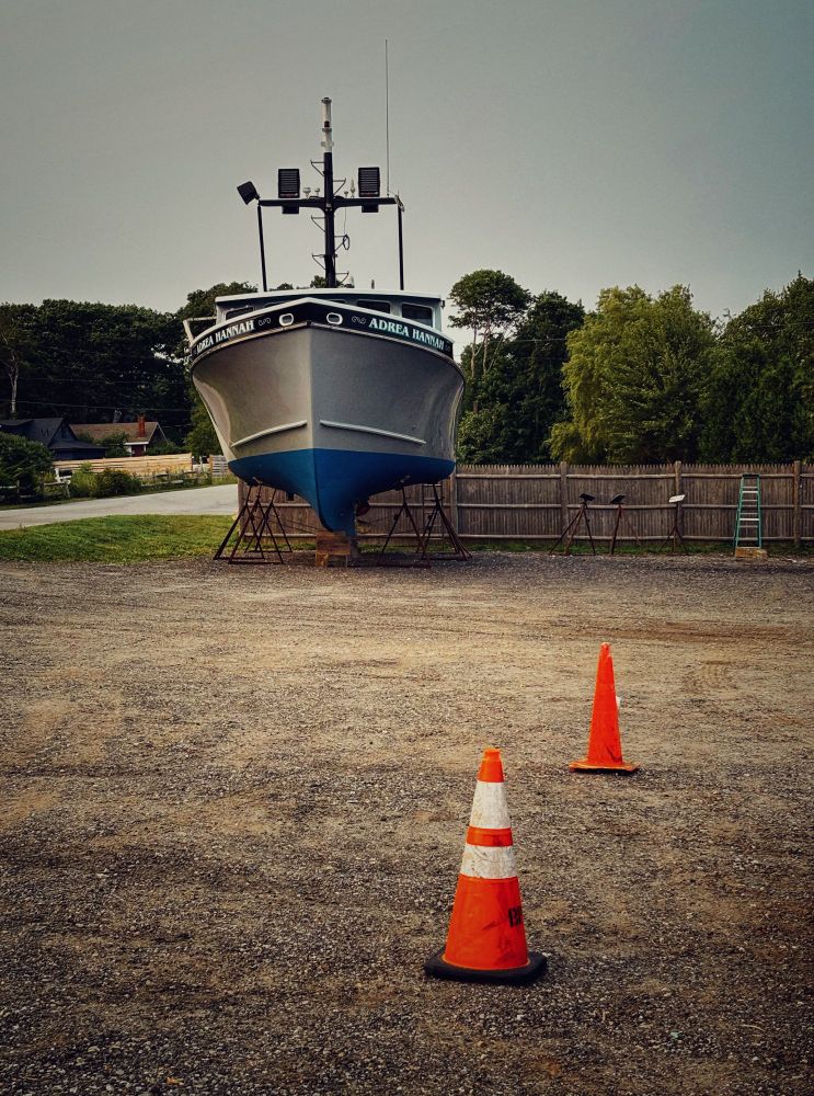 A photo of a lobster boat, facing the camera, hoisted up on pilings in a gravel parking lot. The boat hull has a blue bottom and grey top side, and the name "Adrea Hannah" is written along the gunwale. Two orange road cones in close proximity to each other occupy the bottom right foreground. 