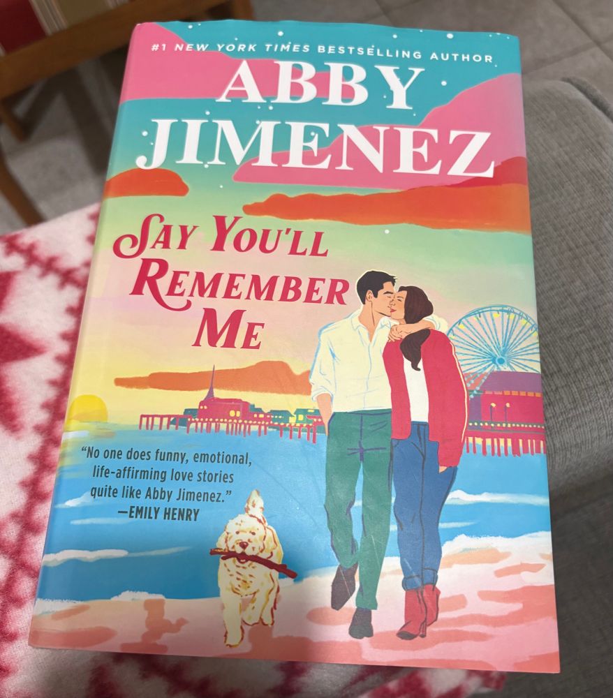 A copy of Abby Jimenez’s book, “Say You’ll Remember Me.” It has bright, pastel colours and artwork. A man and a woman are kissing and walking along a beach with the Santa Monica Pier in the distance. A smaller, fluffy white dog, Jake From State Farm is walking alongside them with a stick in his mouth.