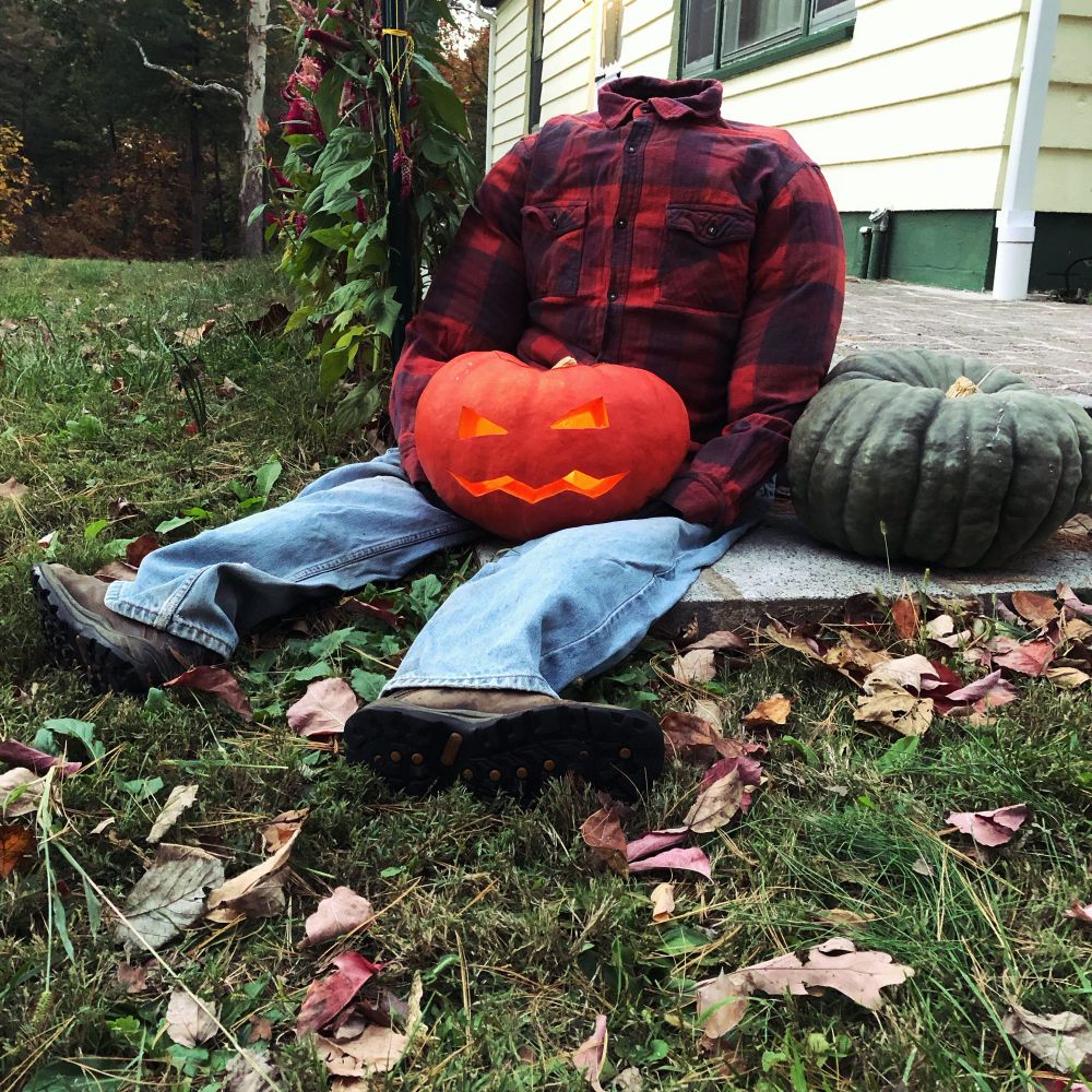 Headless guy in an orange and purple flannel shirt and jeans sits holding a jack-o-lantern