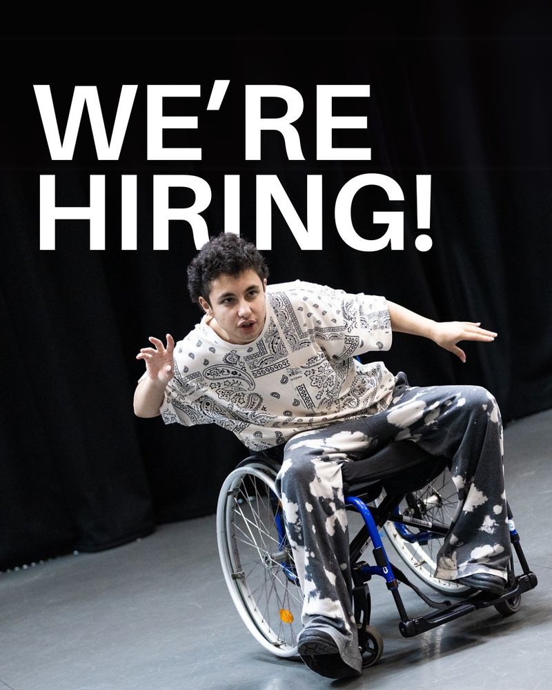 Sam, a mixed-race young man with light skin, dark curly hair, brown eyes and a slim build in his manual wheelchair rehearses Just Short of Saintly. He is wearing a white t shirt with a paisley pattern and grey and white trousers. He leans to his right with arms raised. White text is overlaid reading "we're hiring"