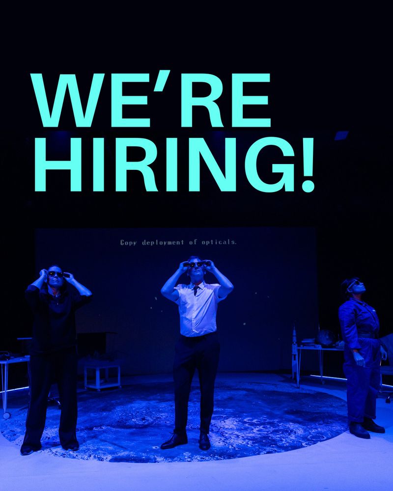 Production shot of The Acts. Anna, a freckled white woman with long brown wavy hair, Matthew, a white male, with light brown hair with a few greys, and Peyvand, an olive skinned woman with a black bob cut and septum piercing wearing a blue cover-all, perform on a stage washed with blue light, they all look up wearing dark sunglasses. Text digitally imposed reads "We're hiring!"