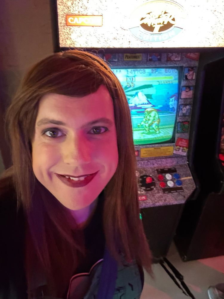 Amy, a person with feminine gender presentation, standing in front of a Street Fighter II arcade machine. She’s got brown hair that goes down just past her shoulders and is wearing a little makeup including a dark red lipstick. She is wearing a green pinafore dress over a black t-shirt, only slightly visible.