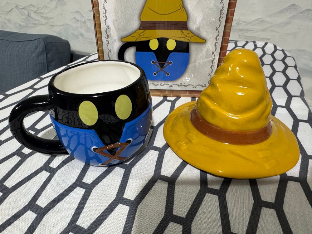 A ceramic mug patterned after Vivi, the black mage from Final Fantasy IX. It has a wide brimmed pointy hat and the body is blue and black with yellow glowing eyes. The hat is off, showing the interior of the mug