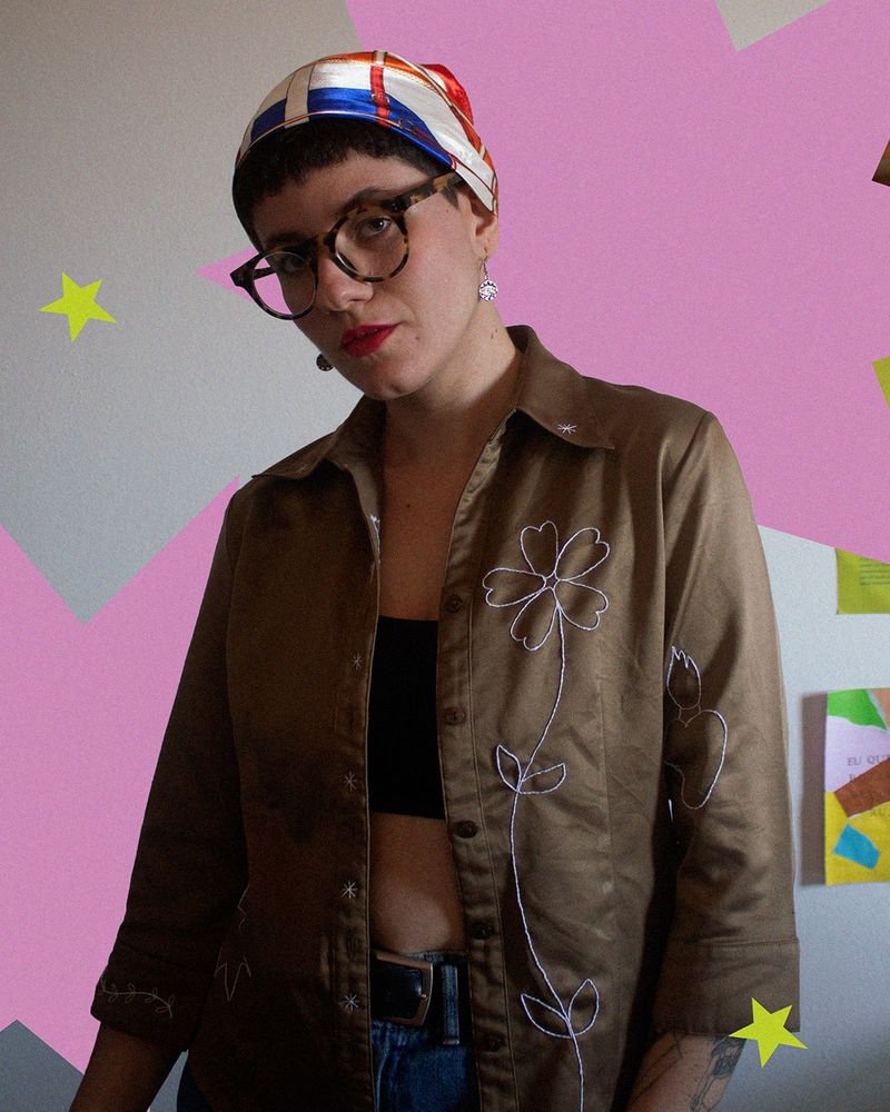 euzinha vestindo a camisa que bordei. tenho cabelo curto e preto e uso um lenço amarrado na cabeça. a foto pega da minha cintura pra cima, estou usando uma camisa marrom meio esverdeada (?) com um tecido meio brilhante e manga 3/4. em uma das mangas da camisa tem o bordado em linhas brancas de um coração saindo fogo. na parte da frente da camisa, do lado esquerdo, tem uma flor bordada com um grande caule e 4 folhinhas. a flor ocupa toda o espaço dessa metade da camisa. do direito, na base da manga, tem um raminho com folhinhas bordadas. e na metade do lado direito, na parte de baixo, tem uma estrela grande bordada.  a camisa está aberta e por baixo uso um top preto e dá pra ver um pouquinho da minha calça jeans e cinto. eu tô olhando diretamente pra câmera, uso um óculos meio arredondado e marrom, batom vermelho, brincos com um pingente redondo com um olho no centro. 