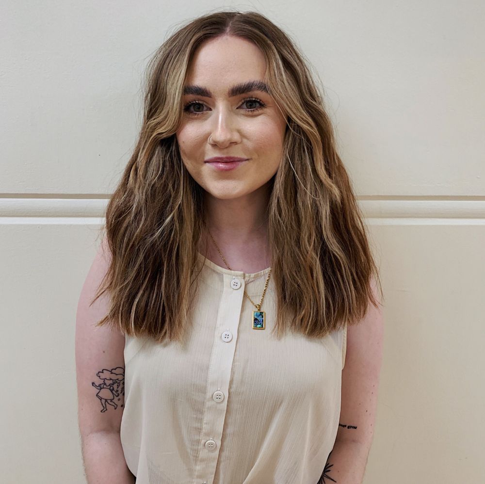 a photo of me, a blonde-haired woman with a cream coloured button-up blouse, tattoos on my arms, and a gold chain necklace with a square blue charm.