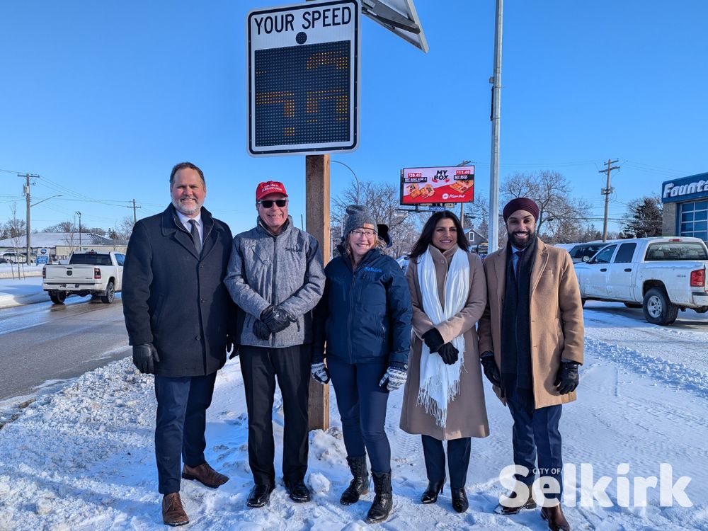 a photo of Minister Matt Wiebe, Mayor Larry Johannson, Deputy Mayor Lorie Fiddler, MPI CEO Satvir Jatana and MLA JD Devgan in front of a digital speed sign on Main Street in Selkirk
