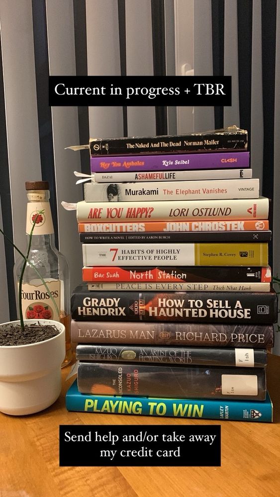 A stack of books on a wood table with a small pencil plant and bottle of whiskey next to the stack. The books are the naked and the dead, hey you assholes, a shameful life, the elephant vanishes, are you happy, boxcutters, how to write a novel, 7 habits of highly effective people, north station, peace is every step, how to sell a haunted house, Lazarus man, an artist of the floating world, the unconsoled, and playing to win
