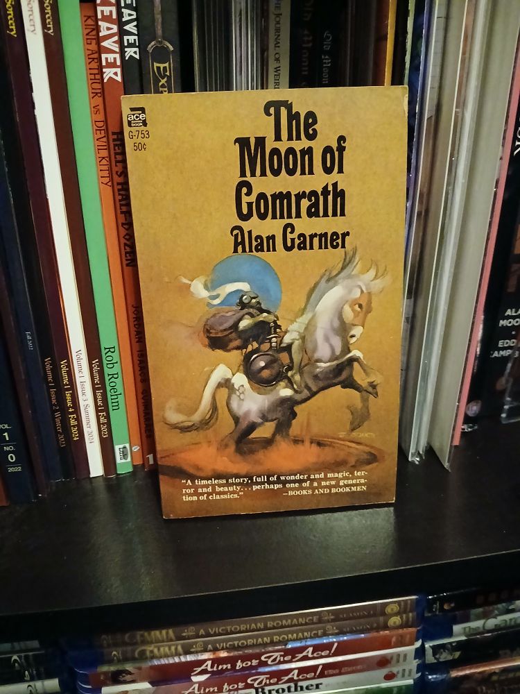 A paperback of 'The Moon of Comrath' by Alan Garner on a bookshelf.