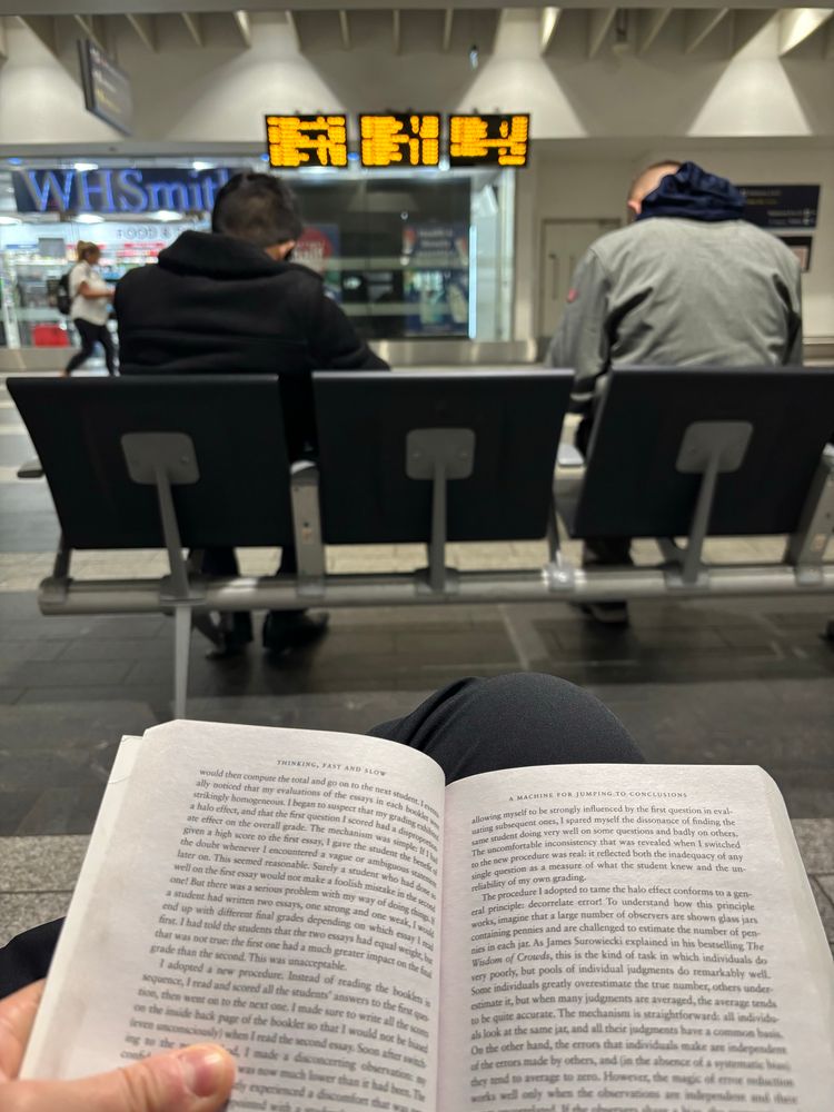 A book in the foreground. A bench with two people on and a departure board in the background.