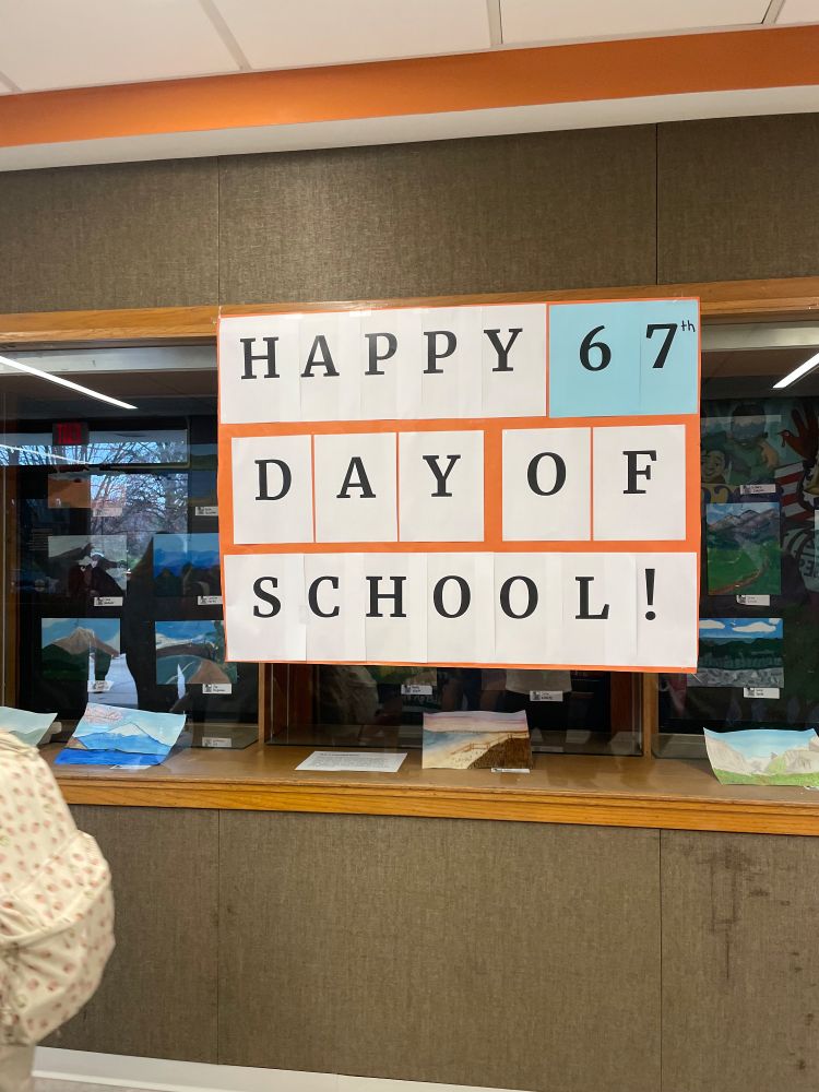 A sign made of 8.5x11" pieces of paper proclaiming "Happy 67th day of school!" The superscript 'th' has been scaled way down to let the hero of the modern child, the number 67, shine. It's also on blue paper instead of white to offset on the orange background of the poster board behind it. It's hung on a display case with pictures in it because we are the Shelbyville of the two towns here and the other school wins all the trophies.