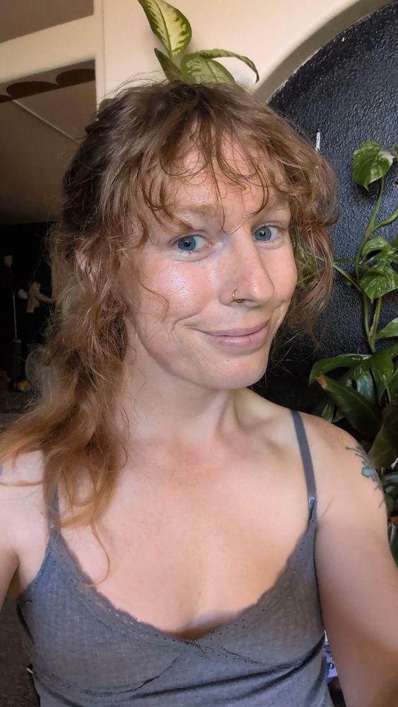 Selfie with messy ponytail and no makeup, in a grey tank top. No glasses on.