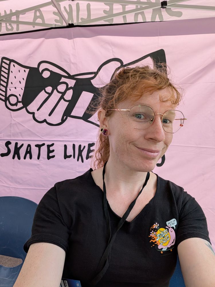 A quick selfie of me in a black shirt in front of the pink skate like a girl banner.
