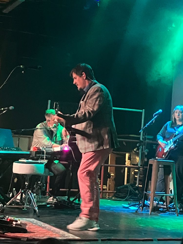 The Mountain Goats perform on stage with John Darnielle center stage holding a guitar and wearing a blazer and salmon colored pants