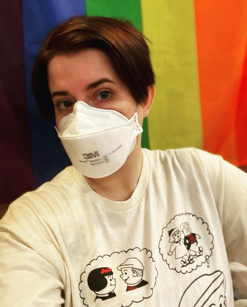 Photo of Whitney sitting in front of a pride flag. They're a white person with short cropped dark brown hair and blue eyes, wearing a cream colored Nancy comic shirt.