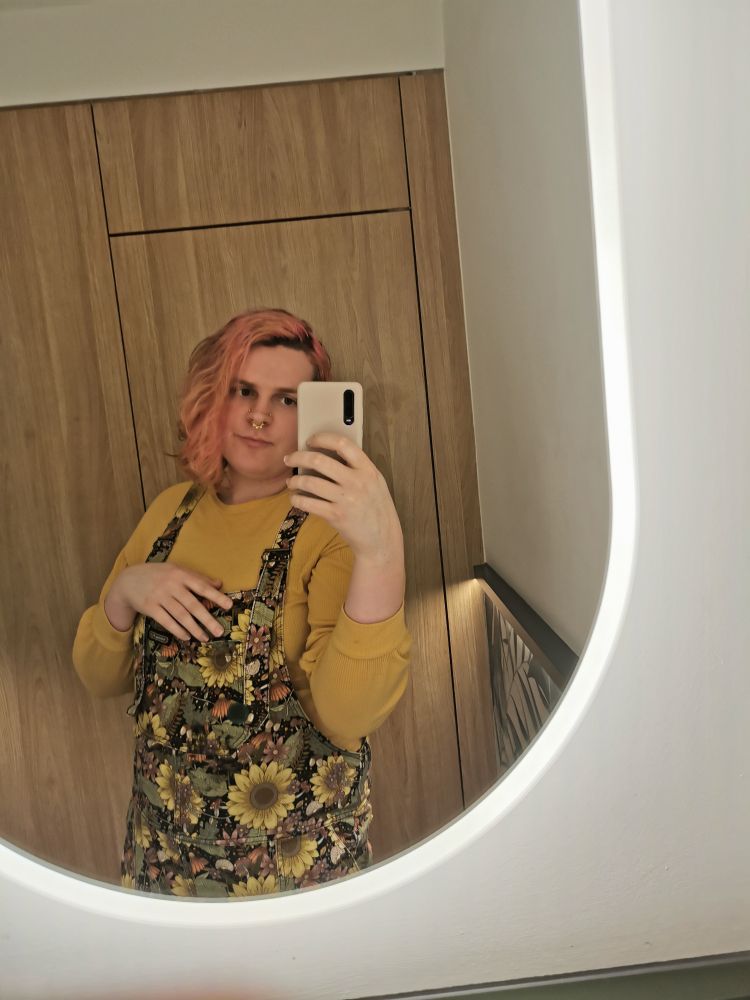 A slightly blurry mid shot of a white woman with pink hair and floral overalls