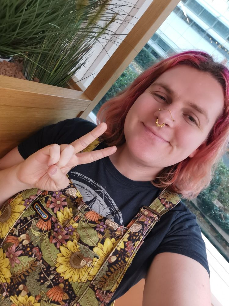 A white woman with pink hair smiling at the camera while wearing floral overalls and a black T-shirt 