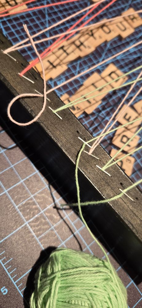 Cropped photo of my studio table, showing a piece of painted black wood, some coloured yarn held by staples, and some wood letter tiles... What could all this be?