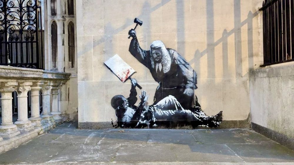 Pintura mural de Bansky en la que puede verse a un juez británico, con toga y peluca,  golpeando con su mazo a un manifestante tirado en el suelo y portando una pequeña pancarta. 