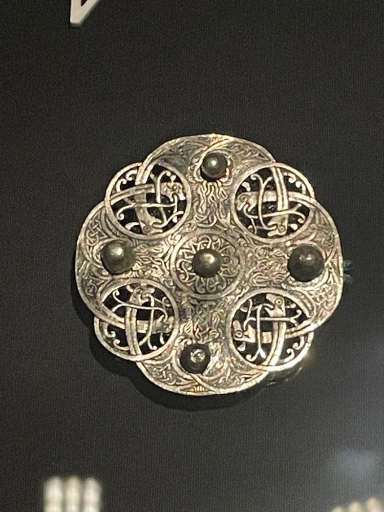 In intricate silver circle brooch with intertwined lines and black silver inlay. There are four circles on the edges in which the interwoven lines end in animal heads. 