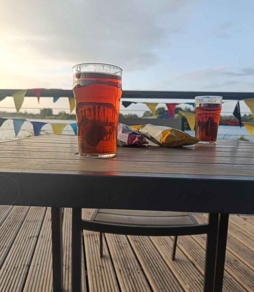 Two pints of chestnut coloured IPA from Rebellion Brewery sit on a table with two packets of post crisps. Bunting adorns the background in front of a broken pale blue sky with dirty clouds.