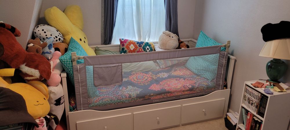 A photo of the same bed from the same perspective.

The length of cloth and mesh above the drawers is now stretched between two poles at the head and foot of the bed, forming a sort of bed rail or fourth short wall to the bed.

Making the small bed functionally an enclosed adult crib.
