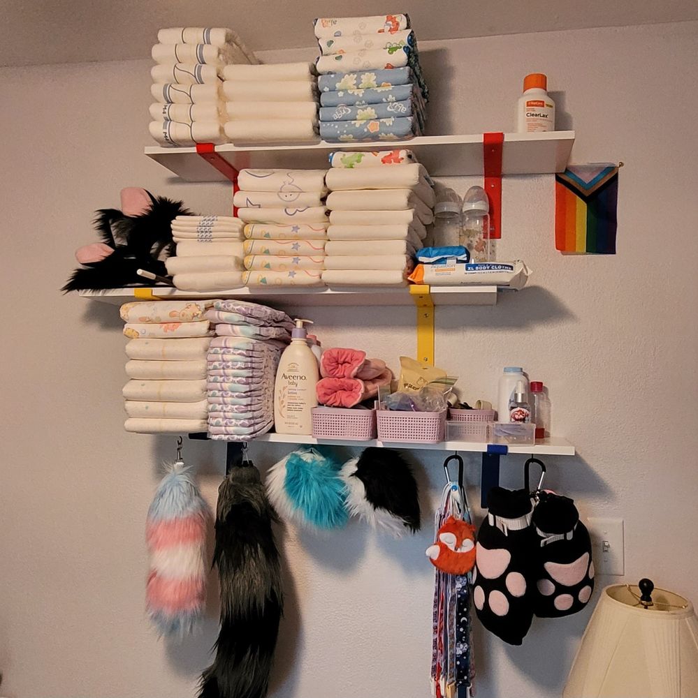 Three staggared shelves stacked vertically. Hanging below the shelves is several tails, pacifier clips, and some plush paw mitts. The bottom shelf has a stack of diapers, pullups, and some small accessories. The other two shelves have more stacks of diapers.
