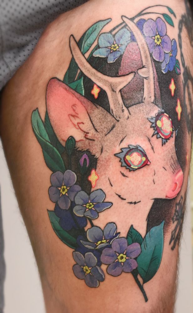 A large thigh tattoo of a deer head with small antlers and three rainbow eyes, surrounded by forget-me-not florals in blue and purple. Little neon stars hover round the deer. The tattoo has a whimsical neotraditional inspired style, with dot shading for the black and grey