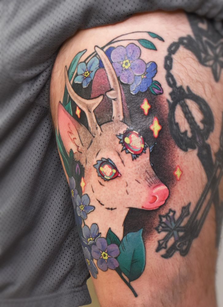 A large thigh tattoo of a deer head with small antlers and three rainbow eyes, surrounded by forget-me-not florals in blue and purple. Little neon stars hover round the deer. The tattoo has a whimsical neotraditional inspired style, with dot shading for the black and grey