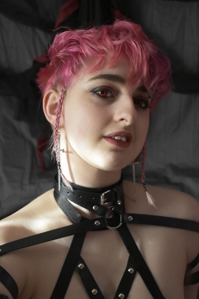 Sylas smiles tilting their head to the side their face lit by natural lighting. They have short pink hair that is curly and tow padawan style braids each framing their face. They wear a black leather strapped harness and chocker studded with silver and sword earrings. Their red eyes accentuated by their messy black eyeshadow.