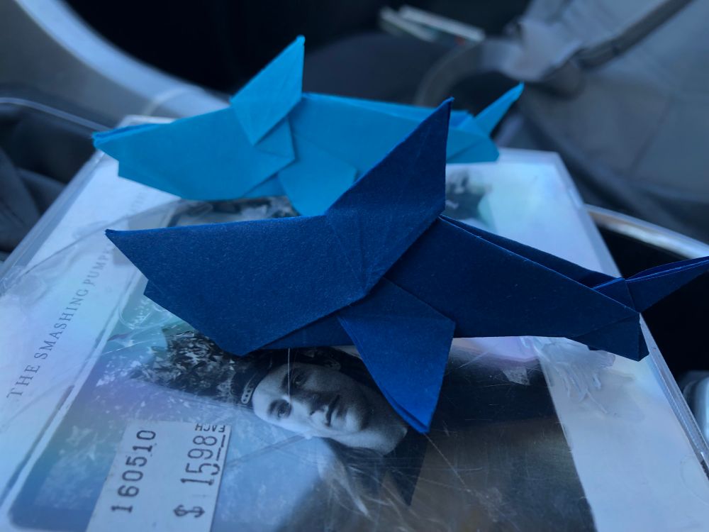 Two origami sharks sit facing left. The closer one is a darker blue while the farther one is light blue. They are both sitting on a “The Smashing Pumpkins Greatest Hits” CD that I got for $15.98.