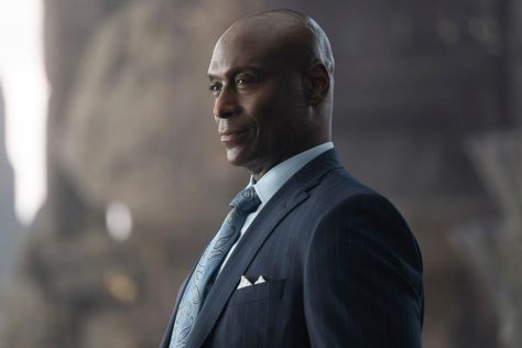 A picture of Lance Reddick. He’s standing and wearing a dark suit with a slate/grey tie and a white button up. 