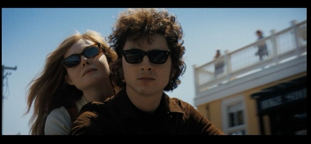 woman with long red hair and sunglasses sitting behind a man with dark curly hair and sunglasses. blue sky behind them, to the right two girls in dresses standing on a white fenced balcony 