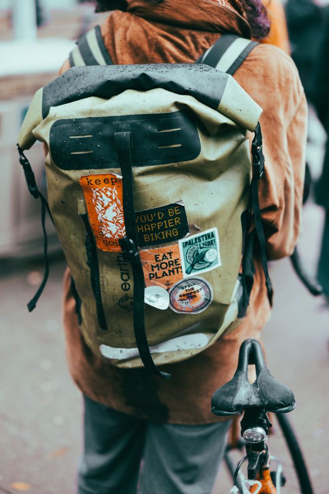 A person seen from the back with a backpack on. One sticker on the back pack reads: You’d be happier biking. 