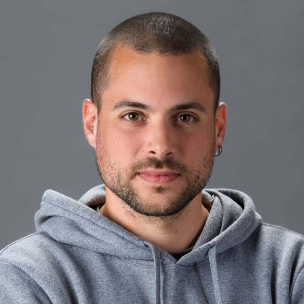 A man with short hair and a beard, wearing a gray hoodie, poses against a gray background. He has light skin, brown eyes, and ear piercings.