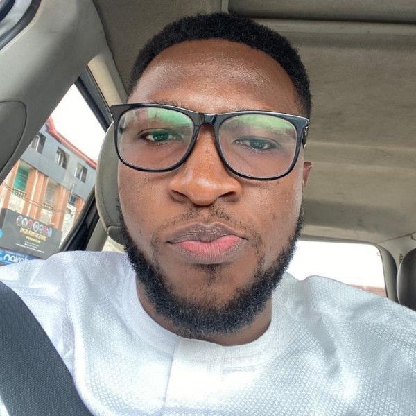 A selfie of a man with dark skin and a short, neatly trimmed afro and beard, wearing black glasses and a white textured shirt. He's sitting in a car, with a glimpse of a street scene visible in the background.