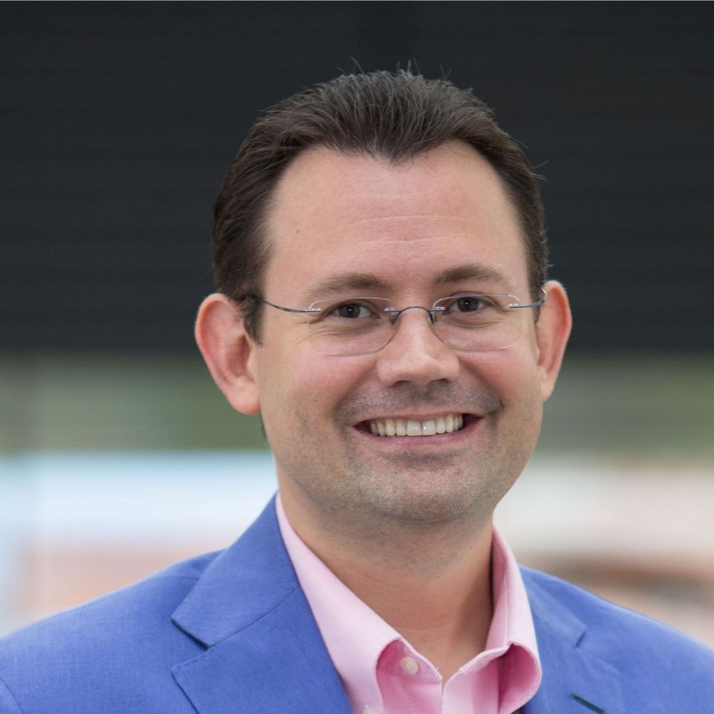 A man smiles wearing rimless glasses, a pink shirt, and a blue blazer. He has short brown hair and fair skin. The background is blurred and dark.