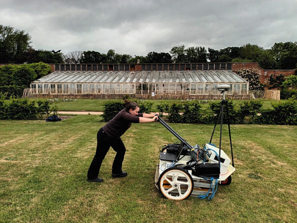 Megan pushing the GPR hand cart with one of the ridge and furrow glass houses visible in the background. The "hot beds" are located under the grass immediately in front of the glass house.
