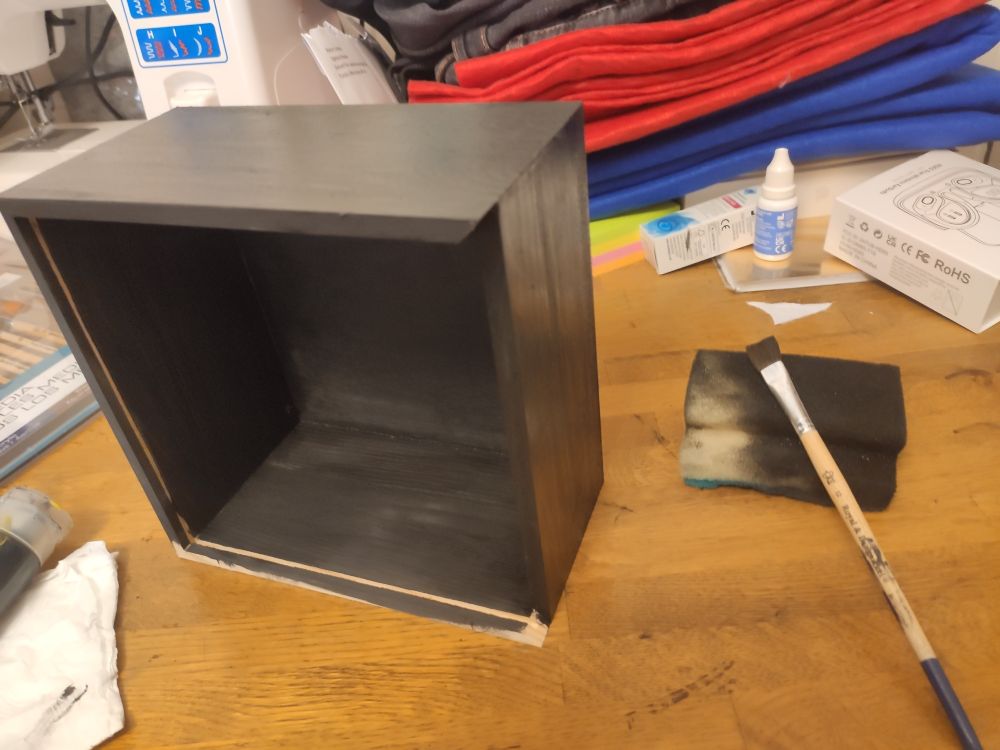A wooden box painted in black with a paint brush a s sponge full of black paint next to it 