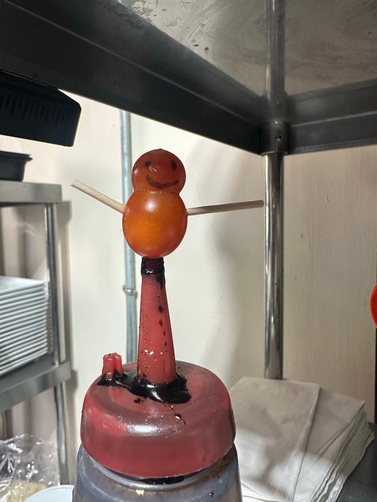 Two little cherry tomatoes fused together with tooth picks sticking out of each side and a smiley face drown on one s
