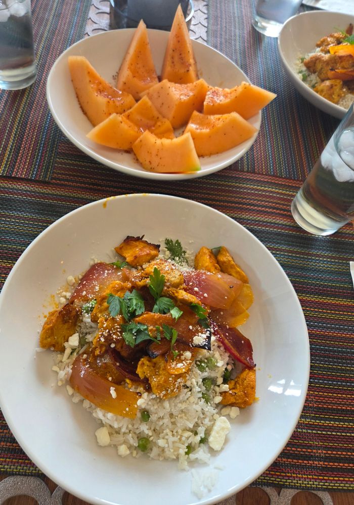 In the foreground a plate of chicken shawarma made on a sheet pan and served over basmati rice and peas. In the background cantaloupe slices topped with Tajin and drizzled with hot honey