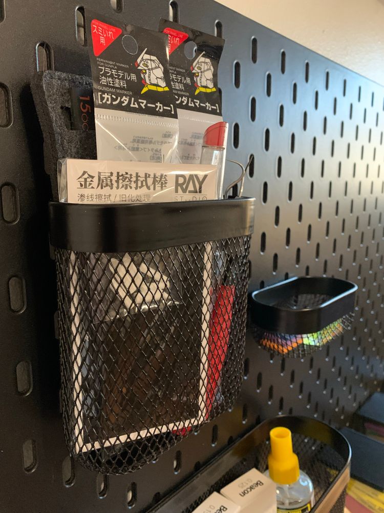 A photo of a black slotted pegboard. There are various baskets and containers hung from the pegboard, filled with various modeling tools. The packaging of the modeling tools contains Chinese and Japanese characters.