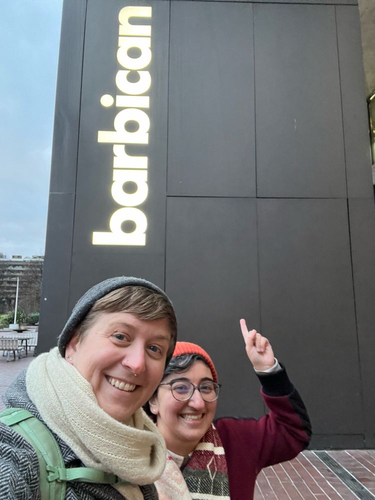 Two people standing infront of a sign that says “barbican”. One is pointing at the sign
