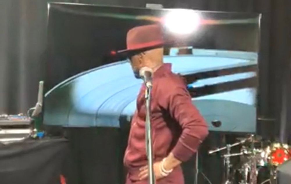 Teddy Riley in all burgundy fitted sweatsuit  and fedora, hands on hips, looking back at tv screen and audio set up during Verzuz with Babyface
