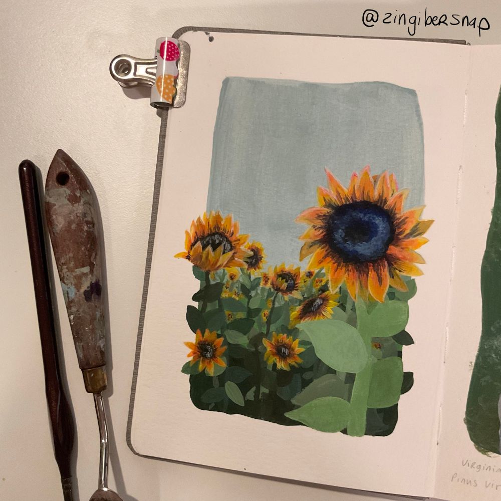 A sketchbook open on a table. The sketchbook’s page shows a gouache painting of some sunflowers in a field. There is a paintbrush and palette knife next to the sketchbook. 